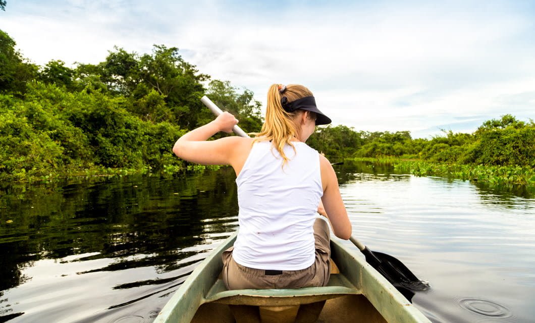 A woman in a white tank top and visor paddles a canoe down a calm, tree-lined river in the Pantanal, surrounded by lush green vegetation under a partly cloudy sky.