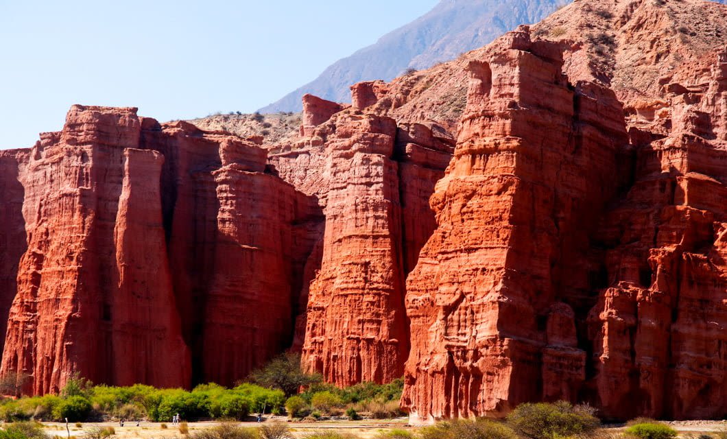 Towering red rock formations rise dramatically against a bright blue sky near Cafayate, with a sparse layer of green vegetation at the base and distant mountains on Salta’s scenic Wine Route in the background.