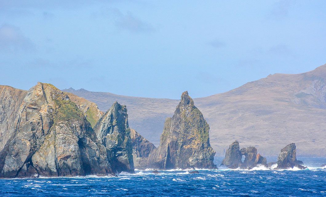 Jagged rocky sea stacks rise from the blue ocean, with waves crashing at their base near the barren coastline of Tierra del Fuego, under a partly cloudy sky—a stunning view often glimpsed on a cruise adventure.