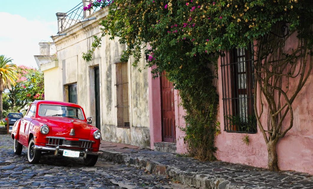 A red vintage car is parked on a cobblestone street in Colonia, Uruguay, beside old, weathered buildings with flowering vines climbing the walls. The scene has a charming, historic atmosphere.