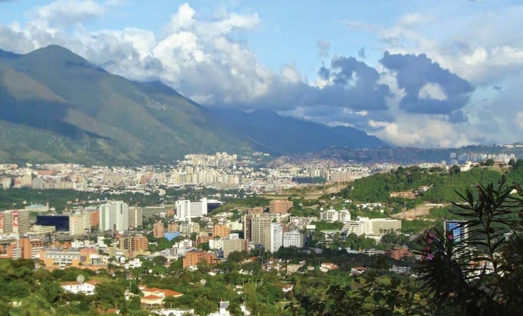 A cityscape with a mix of tall buildings and houses surrounded by green hills, set against mountains under a cloudy blue sky—an iconic scene often featured in Venezuela Highlights for an unforgettable Venezuela Tour.