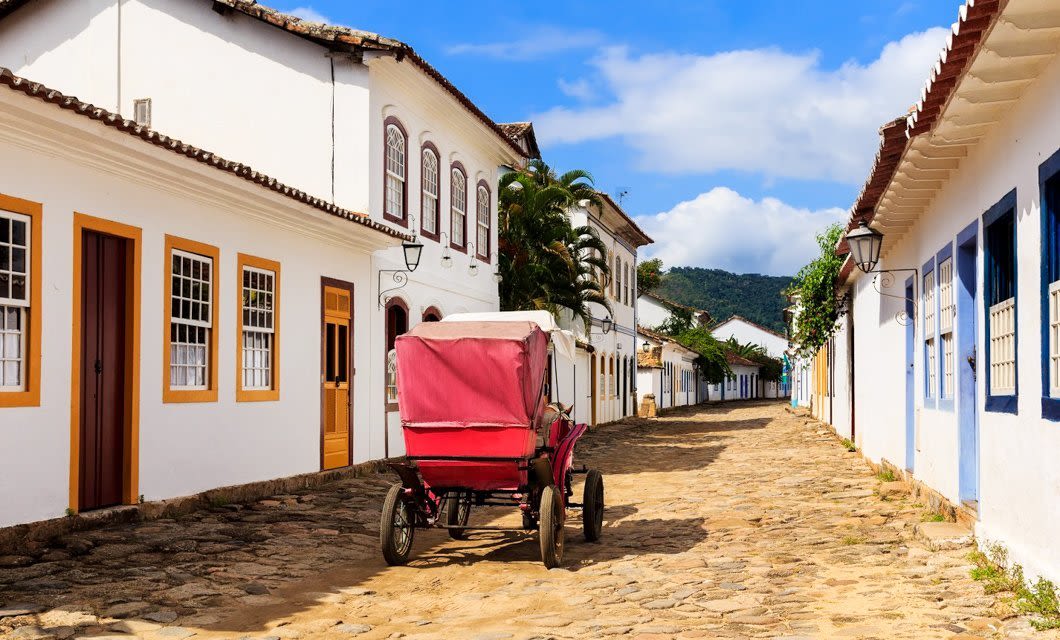 A cobblestone street lined with white colonial buildings, colorful trim, and wooden doors, featuring a red horse-drawn carriage—capturing the charm of the Brazil Gold Route beneath a blue sky with clouds.