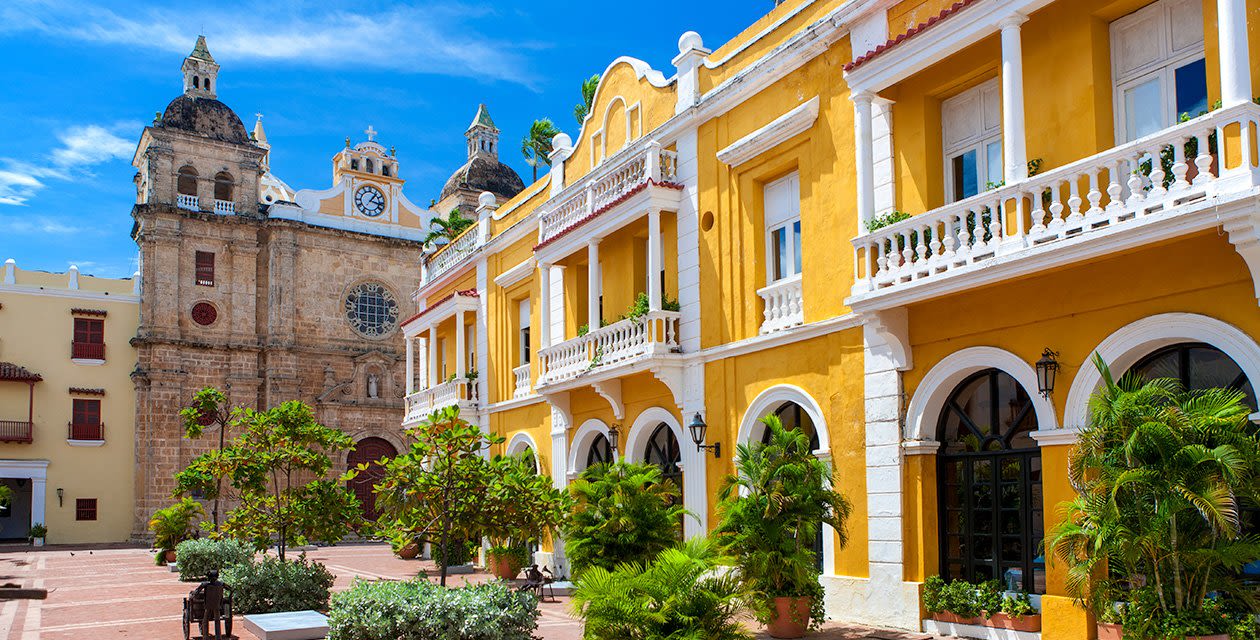 A vibrant yellow colonial building with balconies and arched windows stands next to a historic stone church with a clock tower in Cartagena, under a bright blue sky in a lively plaza featured on the Black History Tour.