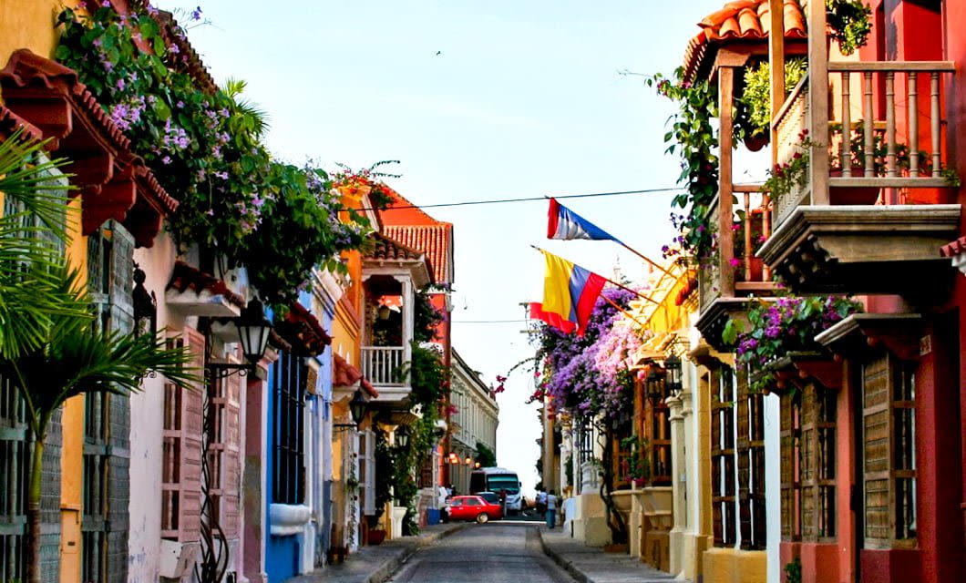 A narrow street lined with colorful colonial buildings, balconies decorated with flowers, and Colombian flags hanging overhead on a sunny day—an inviting scene for travelers enjoying a Colombia golf tour.