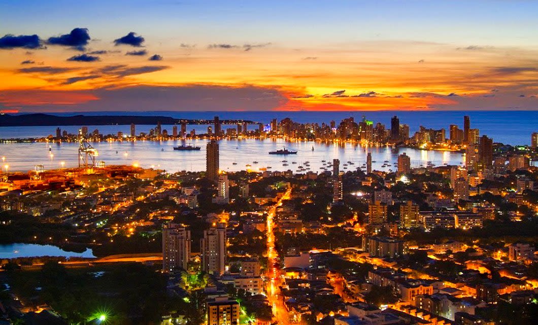 A vibrant city skyline at sunset with tall buildings, glowing lights, and a harbor filled with boats—perfect for a Caribbean tour—set against a colorful sky with orange, yellow, and blue hues reflecting on the water.