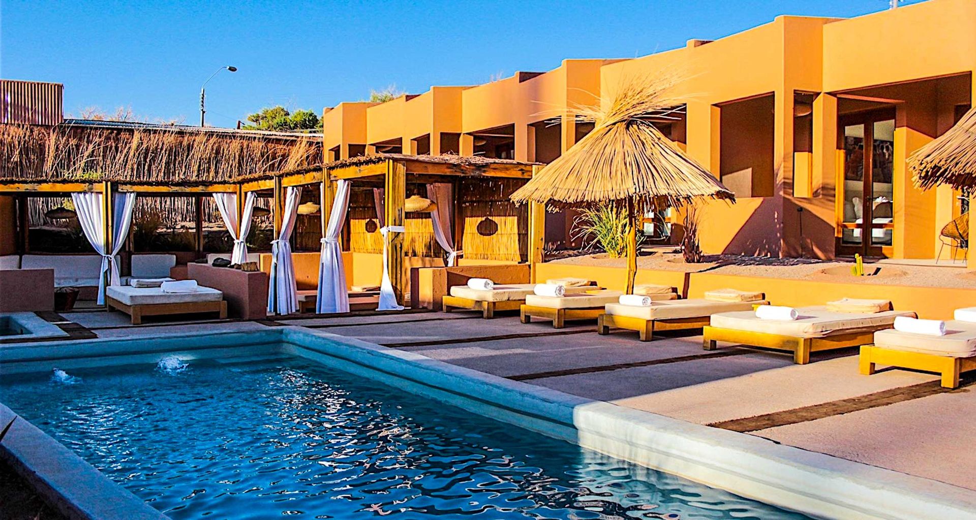 Outdoor pool with clear blue water surrounded by sun loungers with white cushions, cabanas with draped curtains, and thatched umbrellas at Casa Atacama, set against a modern terracotta-colored building under a blue sky.