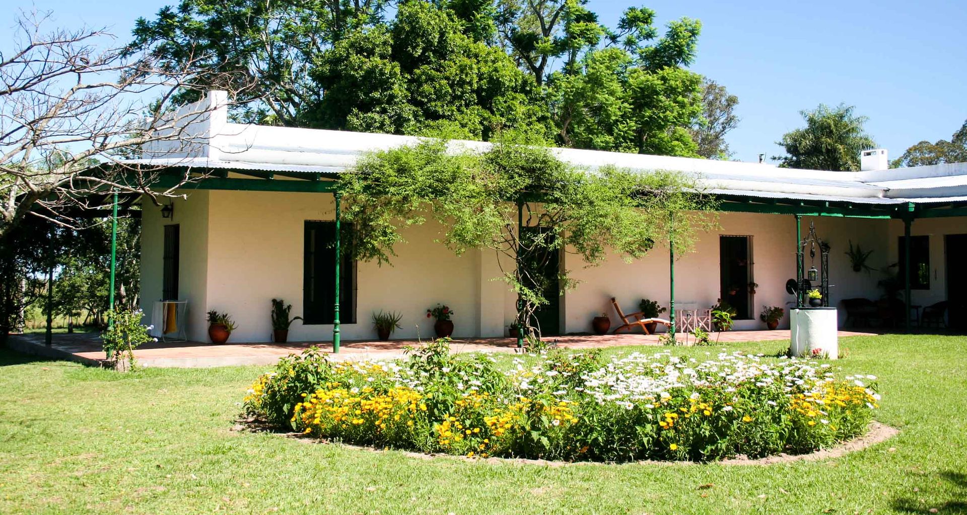 Estancia Buena Vista is a single-story house with a white exterior and green trim, nestled behind a lawn and circular flower bed of yellow and white blooms. Trees, shrubs, and a covered patio area complete the inviting scene.