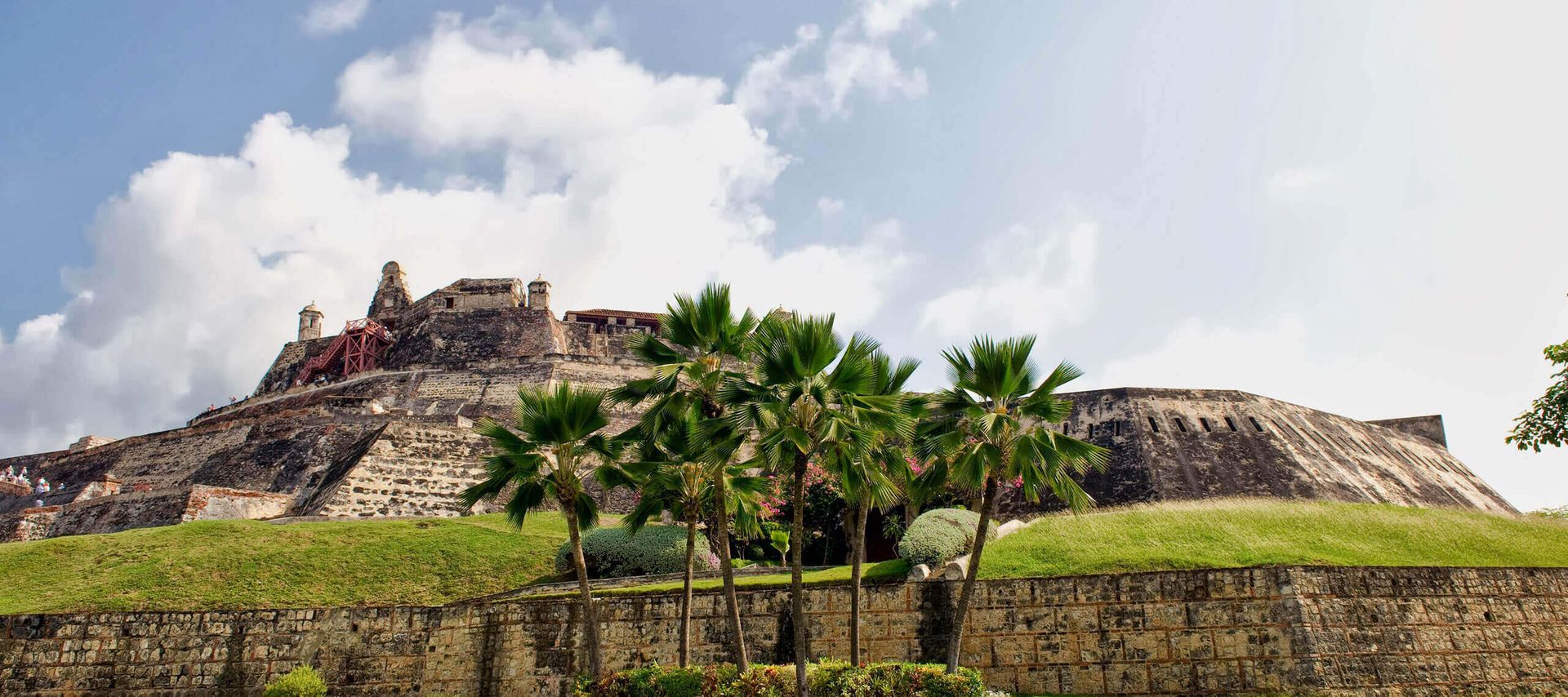 A historic stone fortress with weathered walls sits atop a grassy hill in Cartagena, surrounded by palm trees under a partly cloudy sky—an iconic site often featured on a Black History Tour.