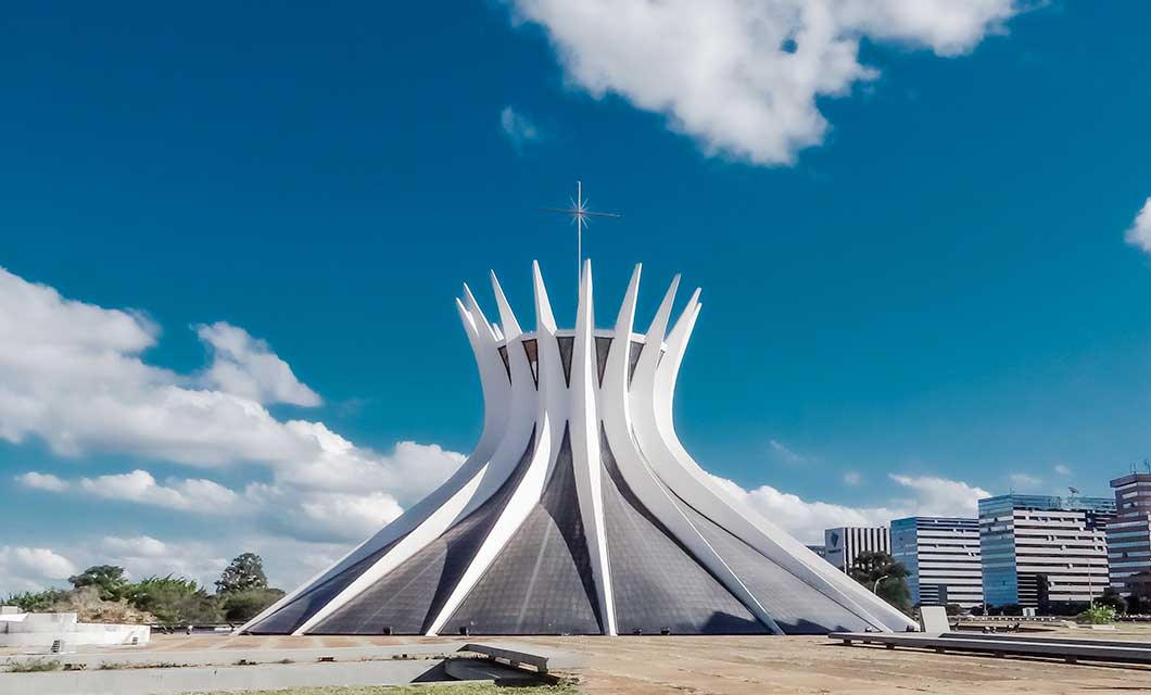 The image shows the Cathedral of Brasília with its distinctive white, crown-like structure under a blue sky, offering a striking contrast to the modern buildings—an architectural gem in Brazil's Cerrado that you can admire during your tour.