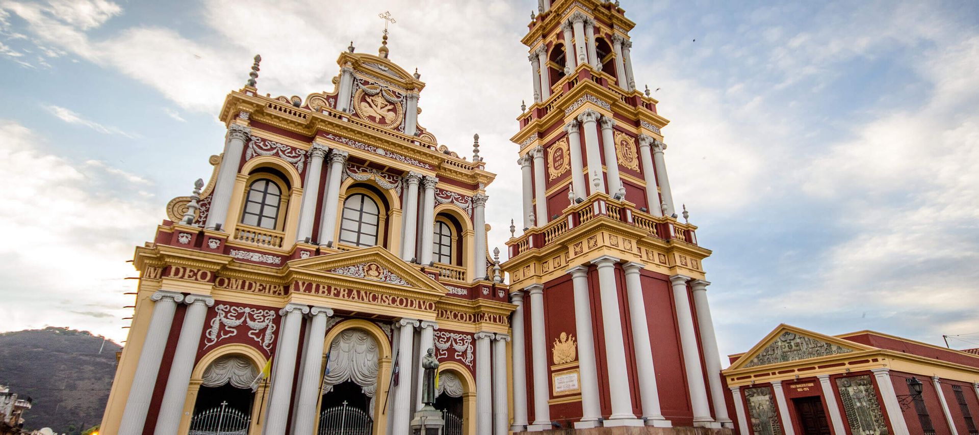 A grand, ornate church with red and gold accents, tall arched windows, and an adjacent clock tower stands under a blue sky. Its intricate architectural details and columns make it a must-see highlight on many Jujuy & Salta travel itineraries.