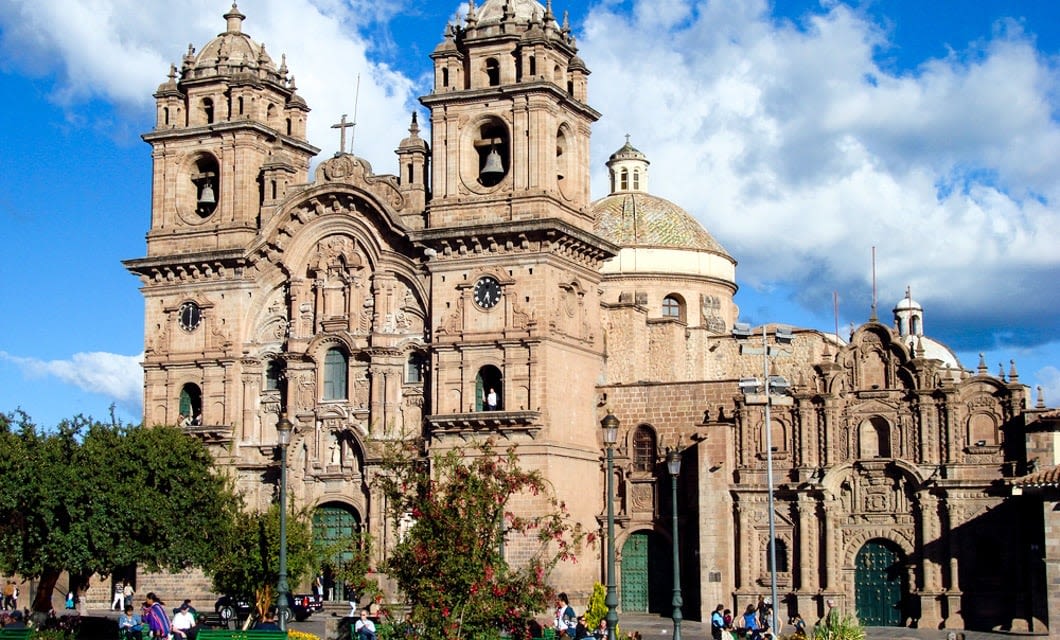 A large, ornate stone cathedral with two towers and a domed roof sits under a blue sky with white clouds. Cariocas walk and gather in front of the building, while trees and plants are visible in the foreground.