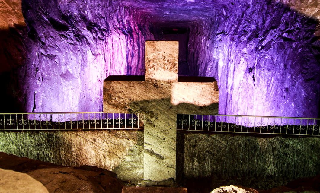 A large stone cross is illuminated by purple and yellow lights inside an underground cave with textured rock walls and a metal railing in front—an iconic sight on a Boyaca, Colombia tour.