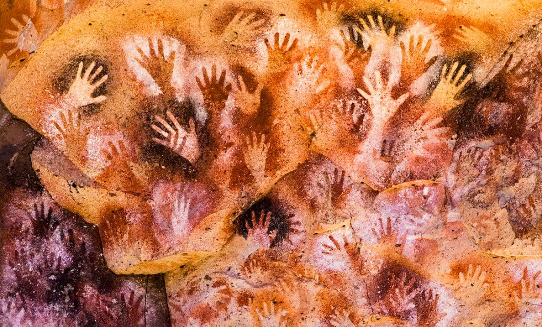 Cave wall covered with stenciled outlines of human hands in various shades of red, brown, and white, overlapping and spread across the textured rock surface—an iconic sight on a Patagonian Route 40 tour.