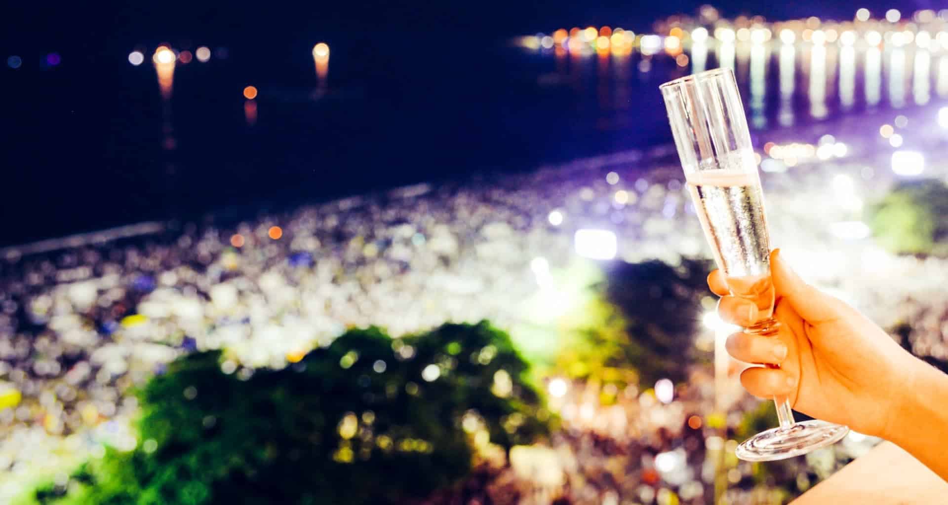 A hand holds a glass of champagne in the foreground, overlooking a brightly lit, crowded beach at night—city lights reflecting on the water—capturing the festive spirit of New Year's in Brazil.