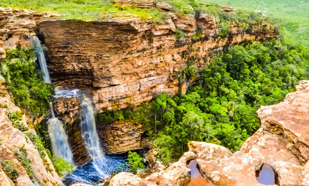 A tall, double waterfall cascades down a rugged, orange-brown cliff into a pool surrounded by lush green forest—an inviting scene for hiking adventures in the stunning Chapada Diamantina landscape.