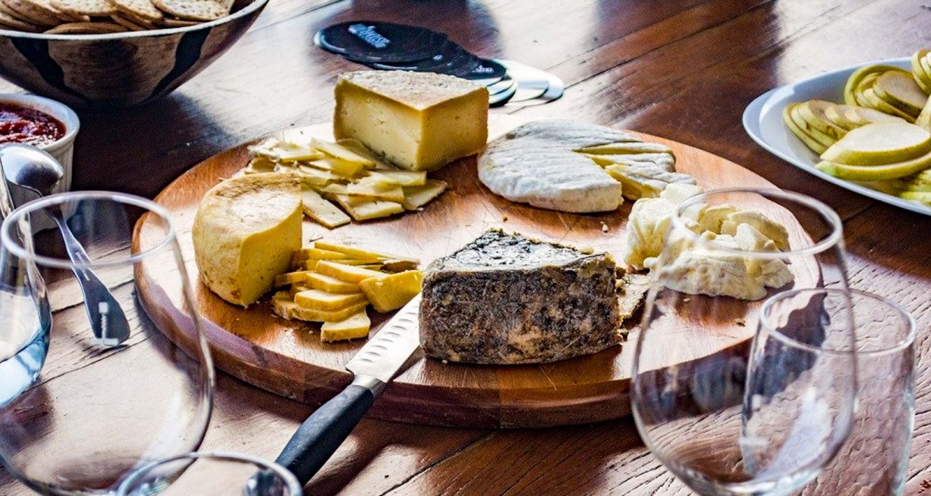 A wooden board with assorted cheeses, sliced cheese, and a knife sits on a table, surrounded by empty wine glasses—perfect for a Brazilian tour or wine tour—alongside crackers, apple slices, and a bowl of jam.