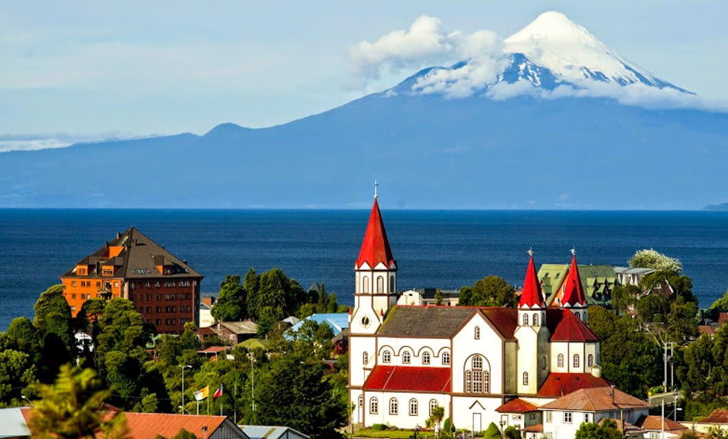 A picturesque Patagonian town with a red-roofed church and buildings by a blue lake, lush greenery, and a snow-capped volcano in the background under a clear sky.