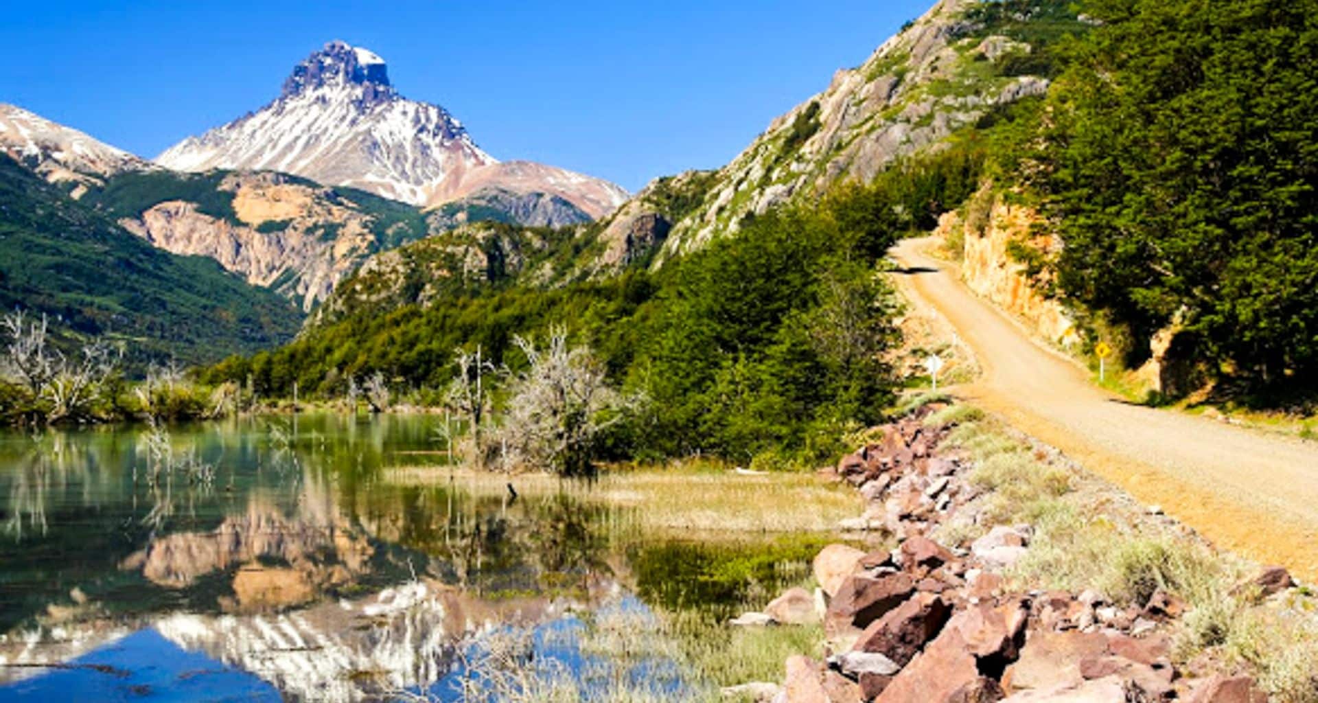 A rugged dirt road runs alongside a reflective lake in Great Northern Patagonia, with dense green trees and rocky hills in the foreground, and a snow-capped mountain peak under a clear blue sky in the background.