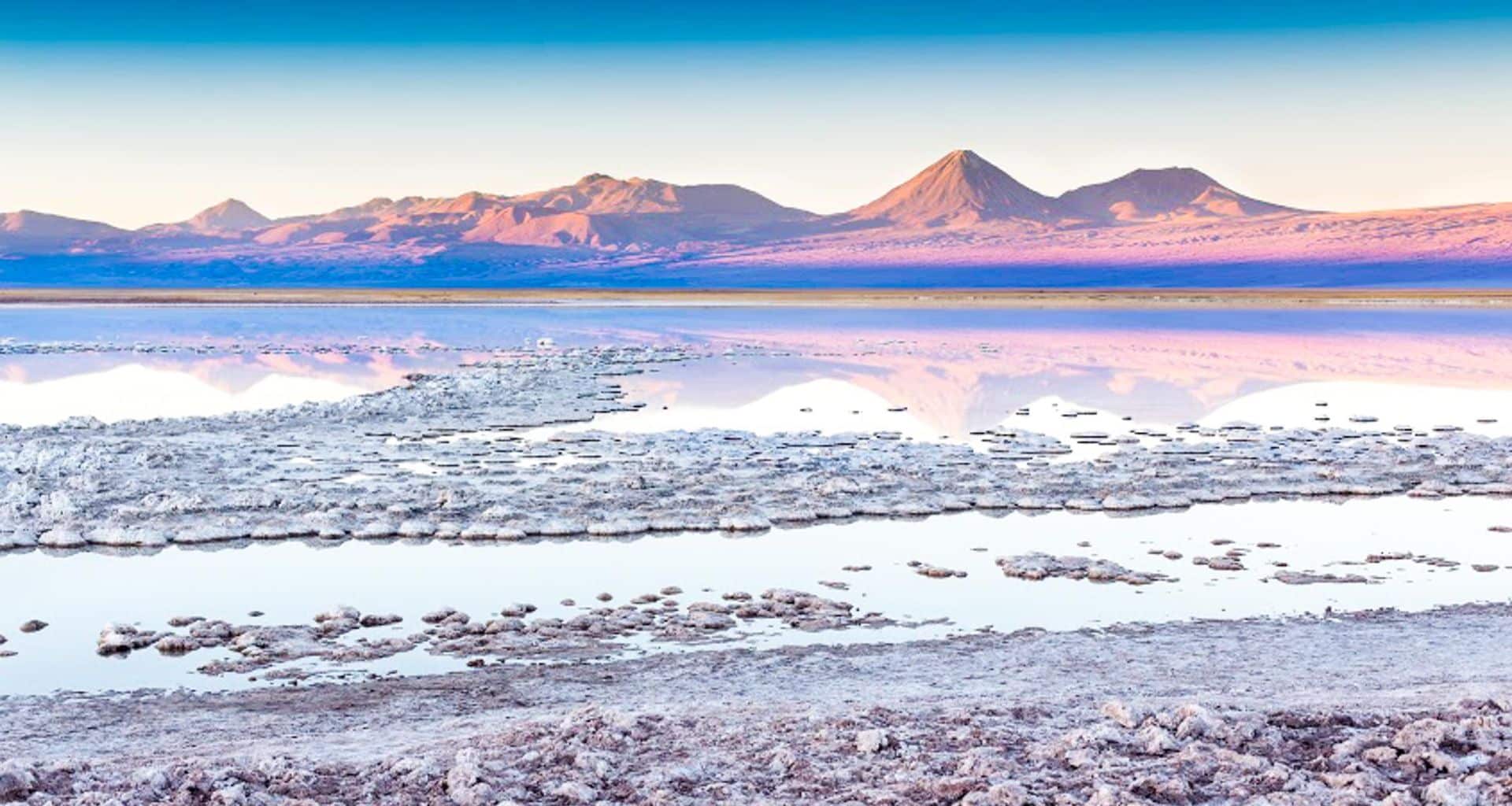 A serene landscape with a salt flat and shallow water reflecting distant pink-hued mountains under a clear blue sky. Perfect for Chile travel, this peaceful scene features textured salt formations in the foreground—one of the hidden highlights of Chile.