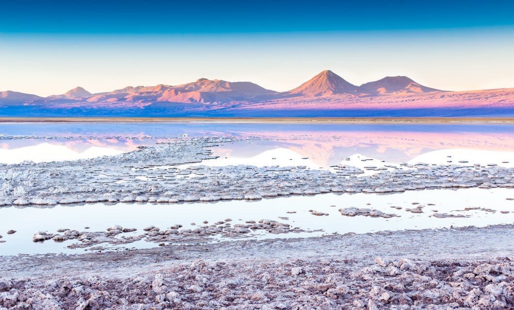 A tranquil salt flat in the Andes with shallow water reflects distant purple and pink mountains under a clear blue sky at sunset or sunrise, capturing the serene essence of the Magic Triangle.