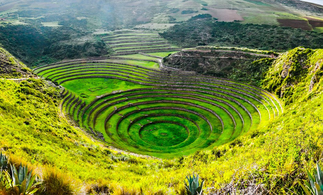 Terraced concentric circular stone structures, known as the Moray ruins, sit in a green valley surrounded by hills and farmland under a bright sky—a highlight of any Peru Train Tour.