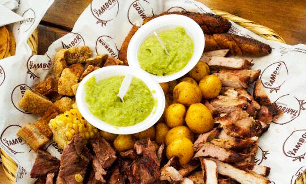 A highlight of the Colombia Gourmet Tour, this platter features assorted grilled meats, corn on the cob, small potatoes, pork cracklings, and two bowls of green sauce, all beautifully arranged on branded parchment paper.