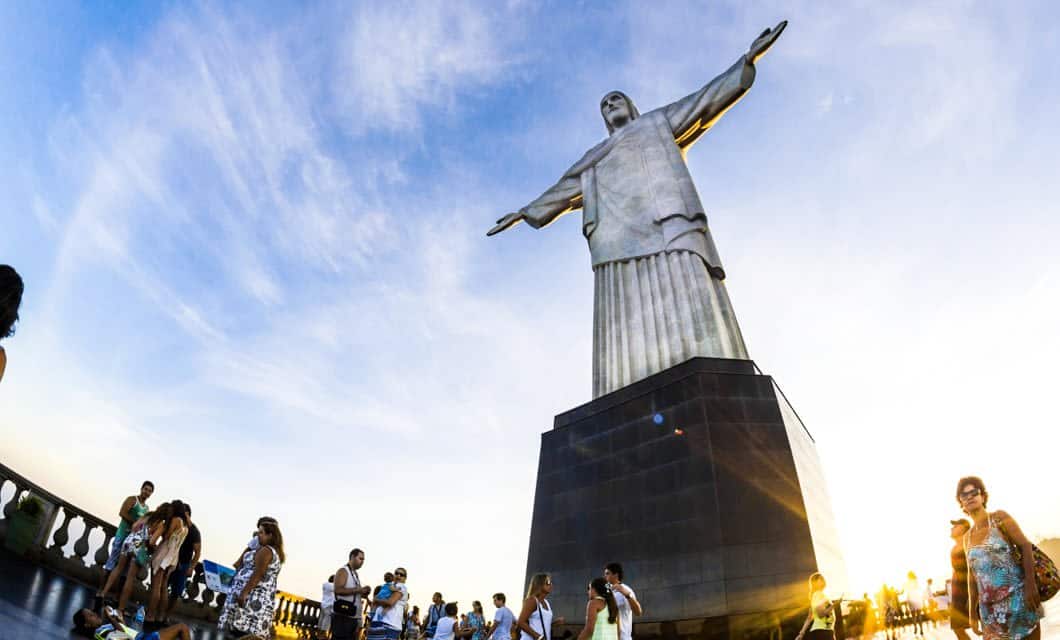 Tourists gather at the base of the Christ the Redeemer statue in Rio de Janeiro, Brazil, under a blue sky with wispy clouds as the sun sets—an unforgettable moment on the Brazil Gold Route or a highlight of any Rio New Years Tour.