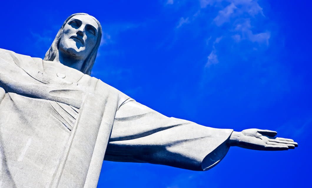 A low-angle view of the Christ the Redeemer statue with outstretched arms against a bright blue sky, capturing the spirit of Rio de Janeiro—a city known for its iconic landmarks and stunning Buzios Beaches.