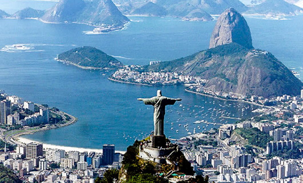 Aerial view of Rio de Janeiro, Brazil, with the Christ the Redeemer statue in the foreground, overlooking the city, beaches, and Sugarloaf Mountain as echoes of samba fill Guanabara Bay in the background.