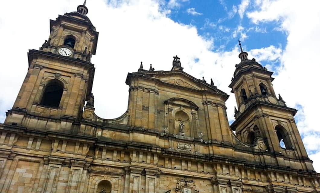 View of a historic stone cathedral in Colombia with two clock towers and statues, set against a partly cloudy blue sky. Architectural details include arches, columns, and decorative carvings—perfect for your Colombia Gourmet tour experience.