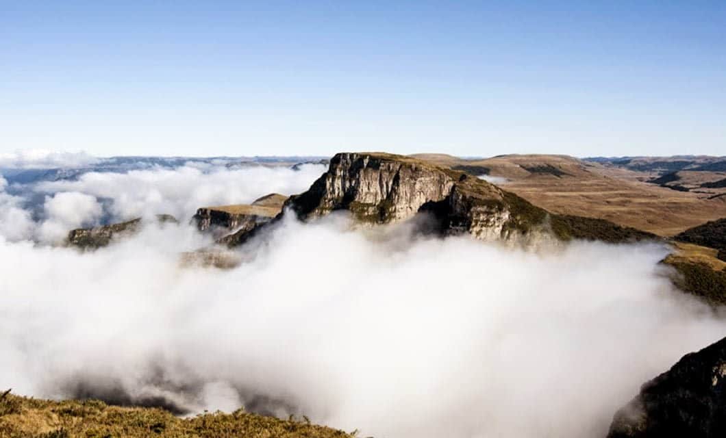 A rocky mountain peak rises above a layer of white clouds in Southern Brazil, with rolling hills and a clear blue sky in the background—perfect for an adventure.