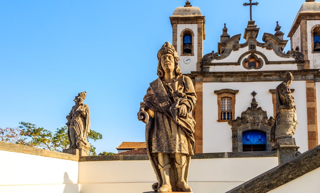 Stone statues of religious figures stand in front of a historic white church with twin bell towers and a cross, under a clear blue sky—an iconic sight along the famed Gold Route on a Brazil Rail Tour.