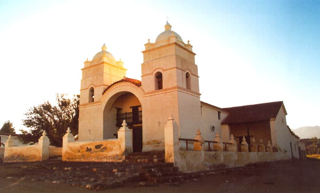 Historic adobe church with two bell towers, arched entrance, and a tiled roof in Cafayate. Nestled along the famed Wine Route in Salta, it is surrounded by a low wall and stone steps, set against a clear blue sky and warm sunlight.
