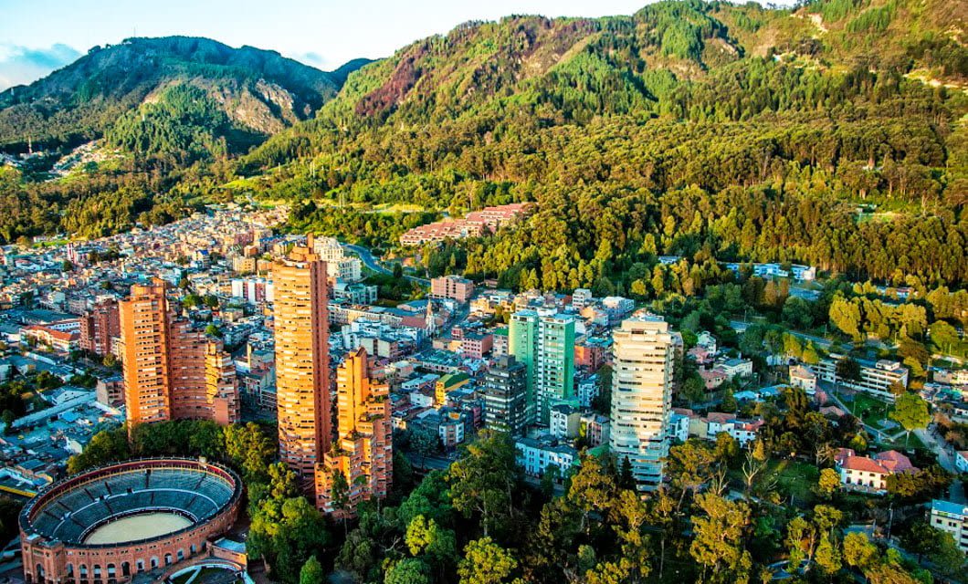 Aerial view of a city with tall buildings, a circular arena, and numerous houses surrounded by lush green forests and mountains—perfect for a Caribbean tour or enjoying authentic Colombia coffee under a partly cloudy sky.