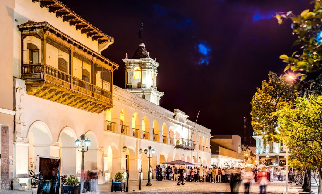 A historic colonial building with arches and a wooden balcony is brightly illuminated at night during the Coast to Coast Tour. A crowd gathers below under a dark sky with scattered clouds, while trees and streetlights frame the lively scene.