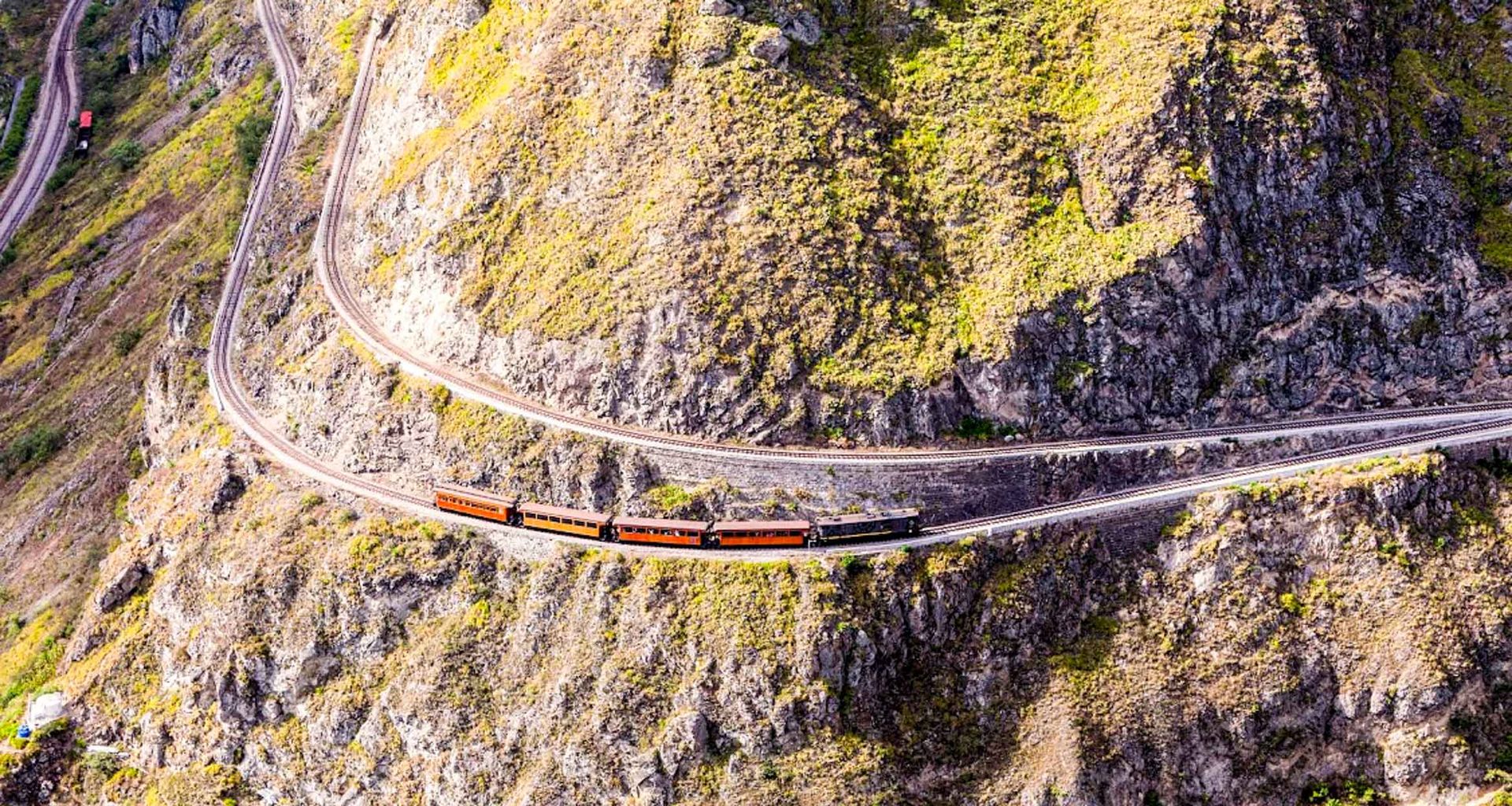 A train with red and black cars, famously known as the Devil's Nose Train, travels along winding tracks on a steep, rocky mountainside covered in sparse vegetation near Quito—a highlight of any Ecuador tour.