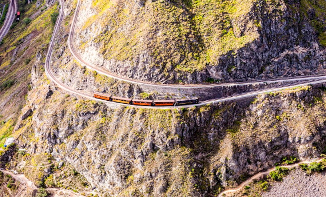 A Tren Crucero with red and black carriages winds along a curving railway track built into the side of a steep, rocky mountain covered with patches of green vegetation in the Avenue of the Volcanoes.