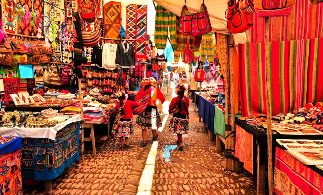 A vibrant outdoor market captures the essence of Peru with colorful textiles, bags, and crafts on display. Two children in traditional clothing walk a cobblestone path between stalls under bright sunlight, reflecting Peruvian culture.