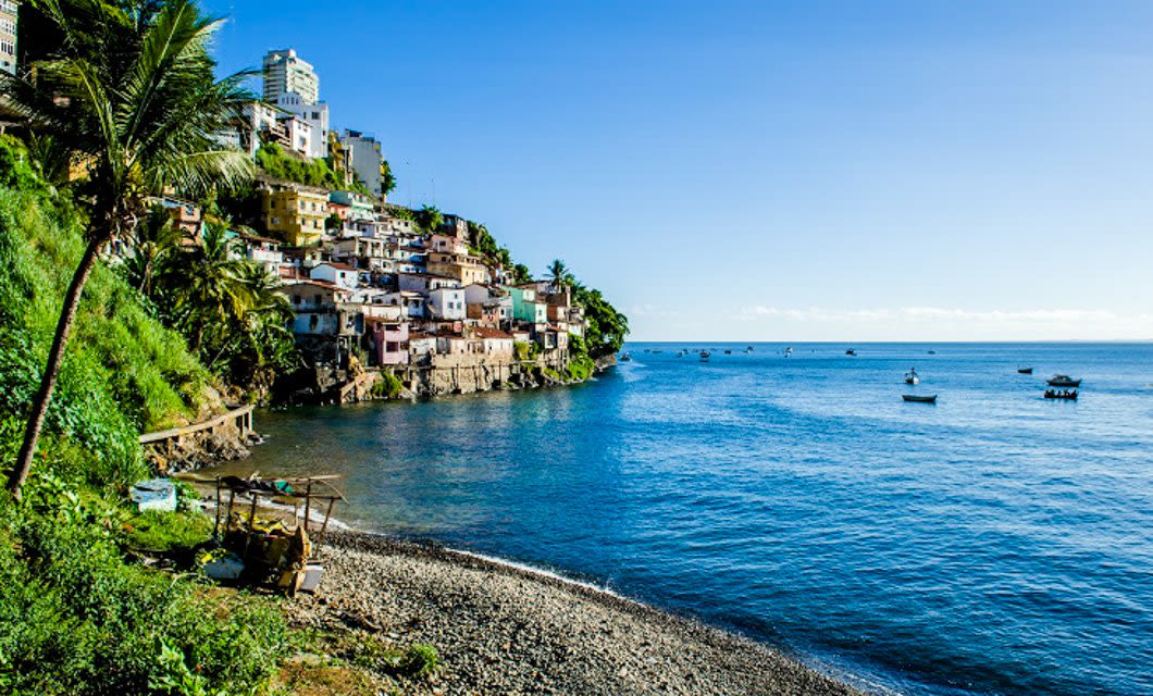 Colorful houses perch on a green hillside overlooking a calm blue bay, with boats scattered on the water and a pebbled shoreline— an inviting scene for hiking adventures reminiscent of Brazil’s Chapada Diamantina under a bright, clear sky.