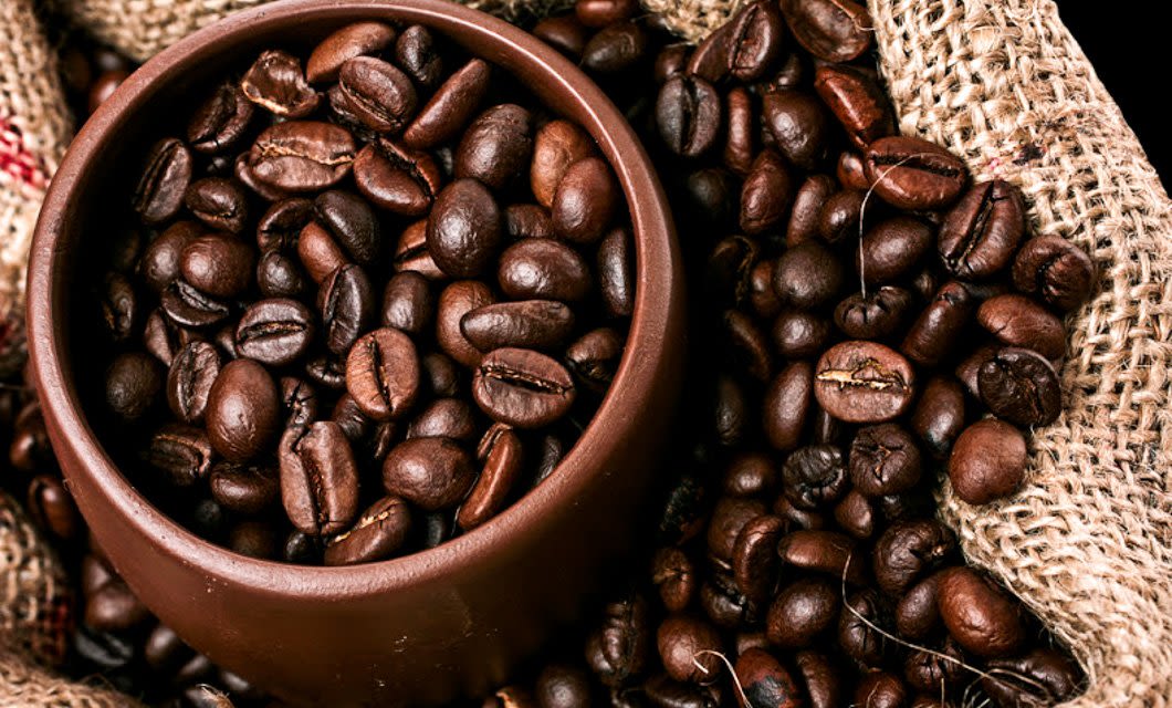 A brown ceramic cup filled with roasted coffee beans rests on burlap fabric, evoking the rich aromas of a Colombia Coffee tour, with more beans scattered around it.