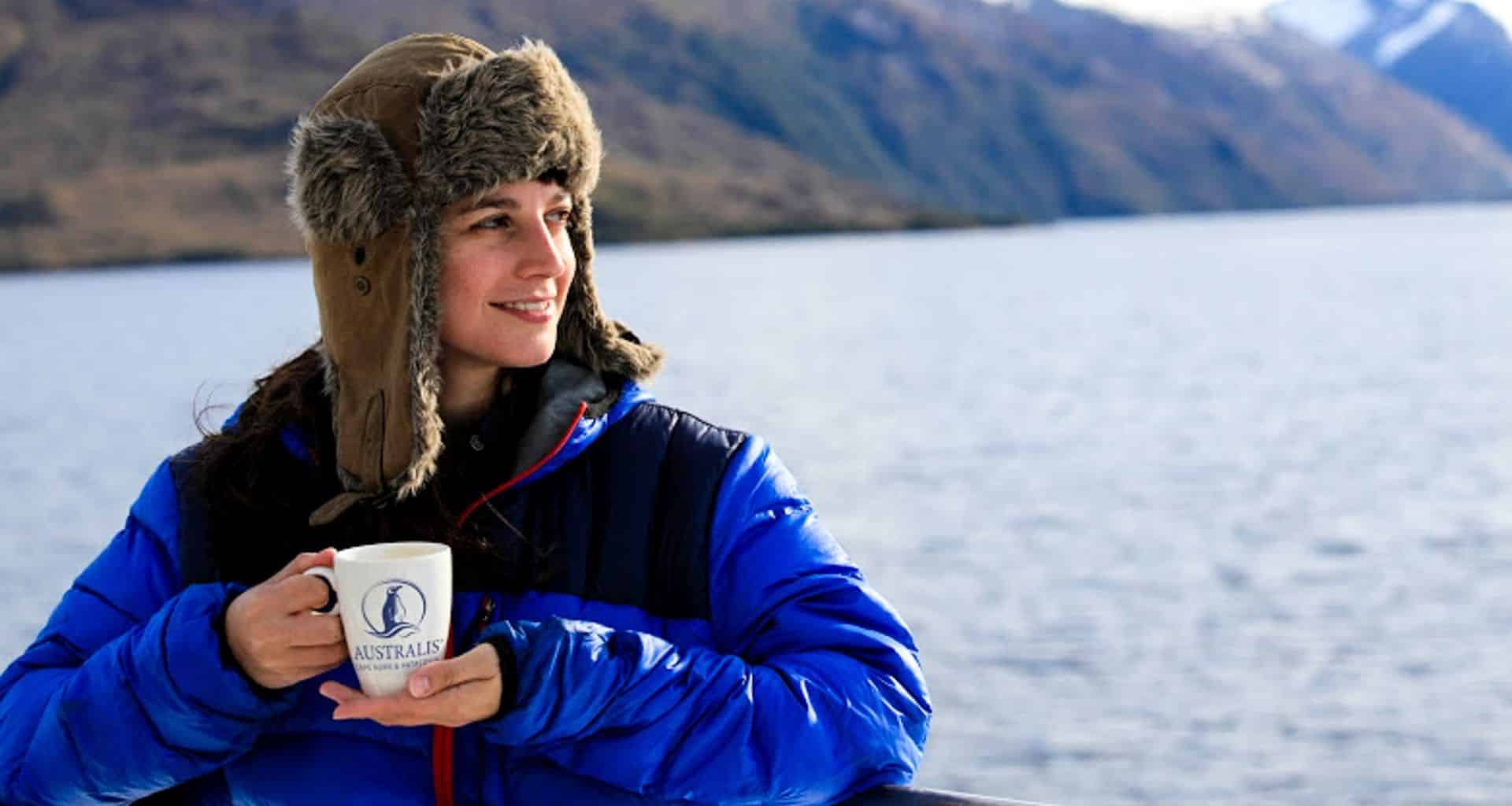 A woman in a blue winter jacket and fur-lined hat holds a mug while smiling and looking to the side, enjoying the stunning views during her Patagonia cruise from Santiago to Buenos Aires, with mountains rising across the water.