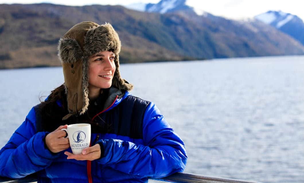 A person in a blue jacket and fur-lined hat smiles while holding a mug, standing by a railing with a lake and snow-capped mountains in the background—perhaps enjoying the crisp air before embarking on a memorable Patagonia Cruise.
