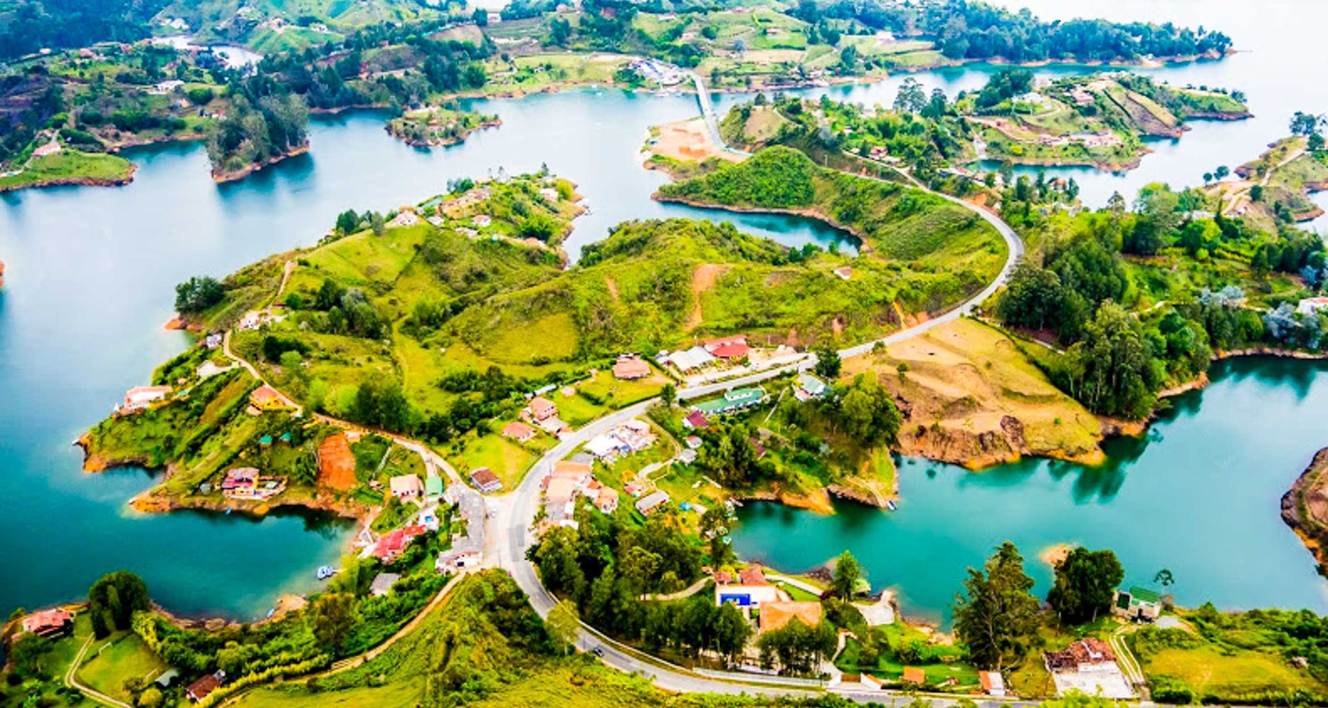 Aerial view of a lush, green landscape with winding roads, small houses, and blue lakes—perfect for a Colombia Botanical Tour—surrounded by rolling hills and scattered trees under a bright sky.