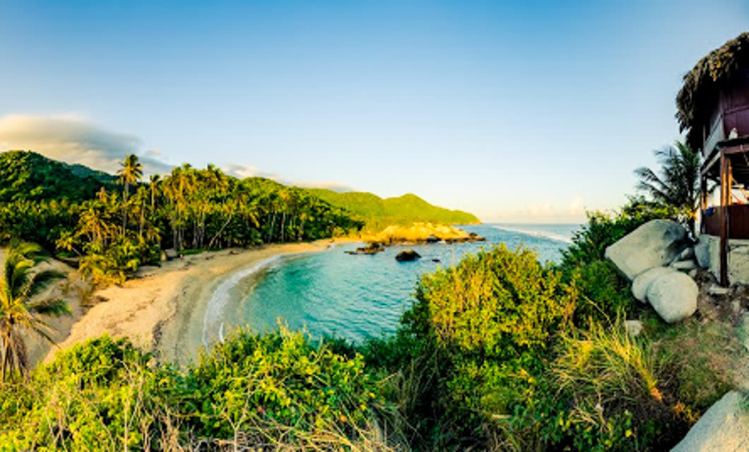 A panoramic view of a tropical Colombian beach with golden sand, turquoise water, lush green palm trees, large rocks, and a thatched-roof hut on the right side under a clear blue sky—perfect for a scenic botanical tour.