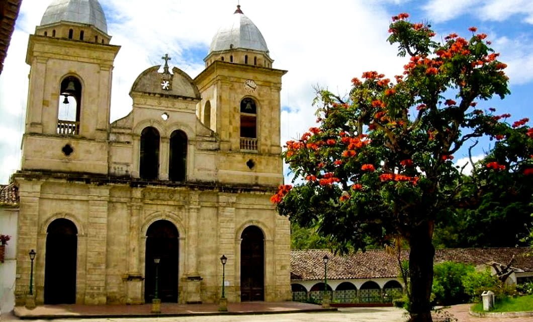 A historic stone church with two bell towers stands beside a vibrant tree with orange-red flowers in Boyaca, Colombia, under a partly cloudy sky. A tiled-roof building and greenery appear in the background, perfect for your next tour.