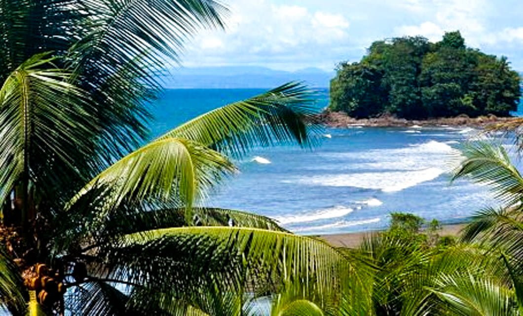 Palm trees frame a view of a tropical beach with gentle waves, blue ocean water, and a small lush island in the distance—an inviting scene perfect for your Colombia Tour along the Caribbean coast under a partly cloudy sky.