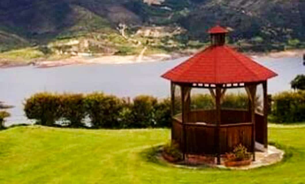 A wooden gazebo with a red roof stands on a well-manicured grassy lawn, overlooking a lake and green hills—the perfect tranquil spot after an exhilarating Colombia golf tour.