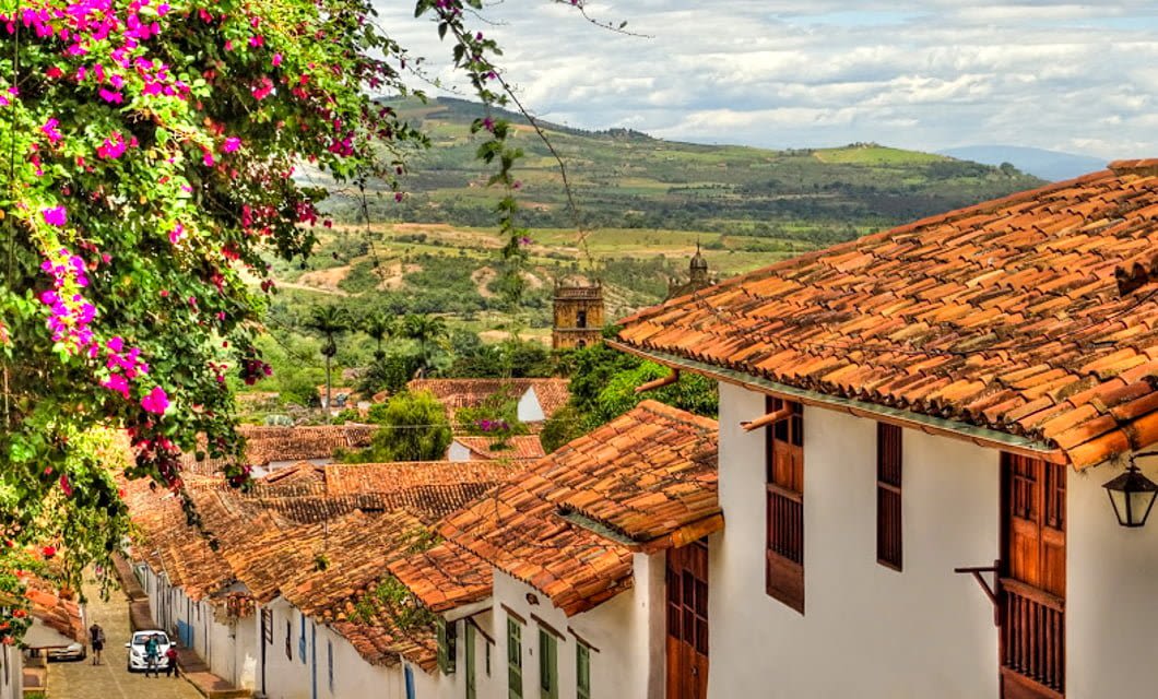 A picturesque village in Boyaca, Colombia, with white colonial-style houses and red-tiled roofs, colorful flowers, and cobblestone streets set against a lush green mountainous landscape under a partly cloudy sky—a perfect spot for your next tour.
