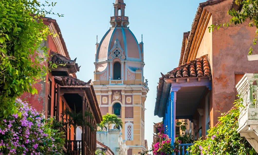 A colorful street in Colombia lined with flowers and colonial buildings leads to a large, ornate dome with a spire under a clear blue sky—the perfect start to a gourmet tour of local flavors and sights.