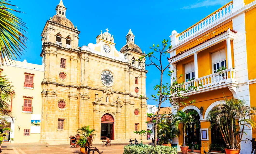 A historic stone church with twin bell towers and a clock stands beside a vibrant yellow colonial building, surrounded by palms and potted plants in a sunny plaza—a perfect stop on your Colombia Coffee or Caribbean Tour.